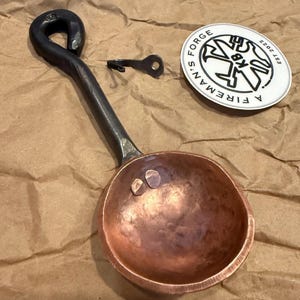 Op de afbeelding: Een handgesmede koperen maatschep met een donker, getextureerd ijzeren handvat met een lusvormig uiteinde. Naast de schep liggen een kleine metalen haak en een ronde witte sticker met de zwarte tekst A FIREMAN'S FORGE EST 2022.