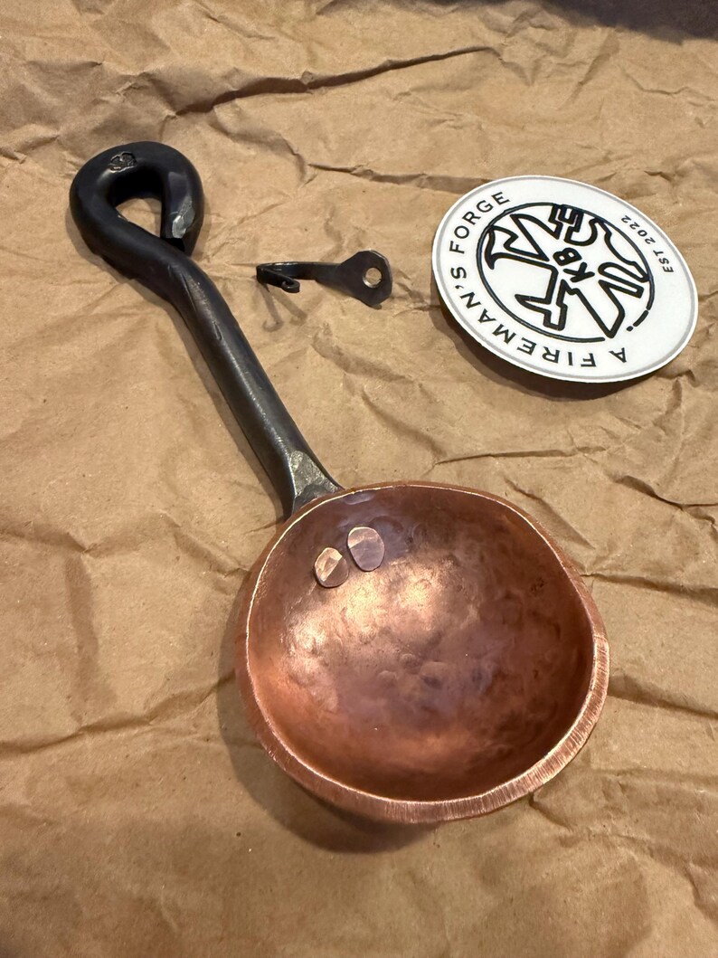 Op de afbeelding: Een handgesmede koperen maatschep met een donker, getextureerd ijzeren handvat met een lusvormig uiteinde. Naast de schep liggen een kleine metalen haak en een ronde witte sticker met de zwarte tekst A FIREMAN'S FORGE EST 2022.