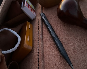 Puede incluir: Una bolsa de cuero marrón con detalles de costura negros. La bolsa está abierta y contiene una pequeña caja de madera, una caja de cerillas, una herramienta de metal negro y dos pipas de madera.