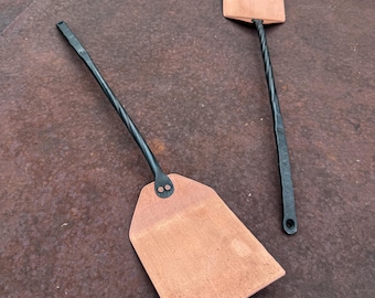 May include: Two spatulas with flat, rectangular copper-colored heads and long, black, twisted metal handles. Each handle is securely attached to the head with two visible rivets. The handles feature a loop at the end for hanging purposes.