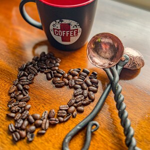 Op de afbeelding: Een zwarte mok met een rode binnenkant en een wit rond logo met het woord COFFEE in rode letters, vergezeld door twee koperkleurige metalen scheppen met gedraaide zwarte handvatten en een hartvormige opstelling van gebrande koffiebonen.