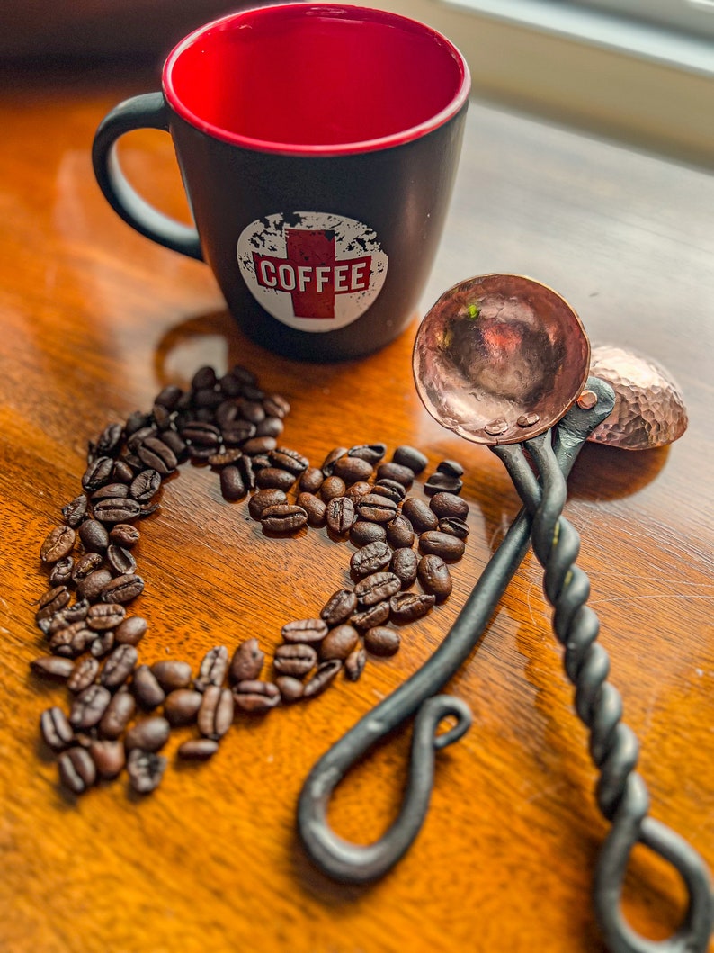 Op de afbeelding: Een zwarte mok met een rode binnenkant en een wit rond logo met het woord COFFEE in rode letters, vergezeld door twee koperkleurige metalen scheppen met gedraaide zwarte handvatten en een hartvormige opstelling van gebrande koffiebonen.
