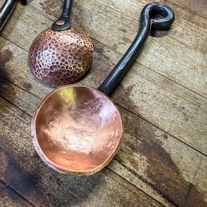 Op de afbeelding: Twee koperen scheppen met ronde, gehamerde kommen en lange, donkere, gesmede metalen handvatten. Elk handvat heeft een gekrulde lus aan het uiteinde. De koperen oppervlakken vertonen een metaalglans met zichtbare inkepingen van het hameren.