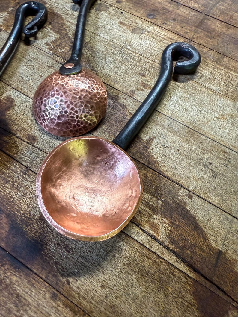 Op de afbeelding: Twee koperen scheppen met ronde, gehamerde kommen en lange, donkere, gesmede metalen handvatten. Elk handvat heeft een gekrulde lus aan het uiteinde. De koperen oppervlakken vertonen een metaalglans met zichtbare inkepingen van het hameren.