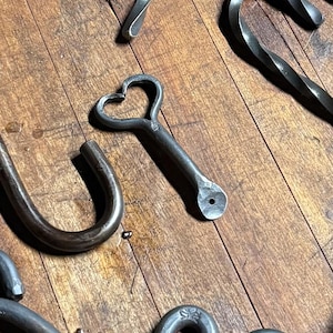 Op de afbeelding: Een verzameling donkergrijze, handgesmede metalen onderdelen, waaronder gedraaide staven, J-vormige haken en een hartvormig element. Verschillende onderdelen hebben platte uiteinden met voorgeboorde gaten voor montage en een ruwe afwerking.