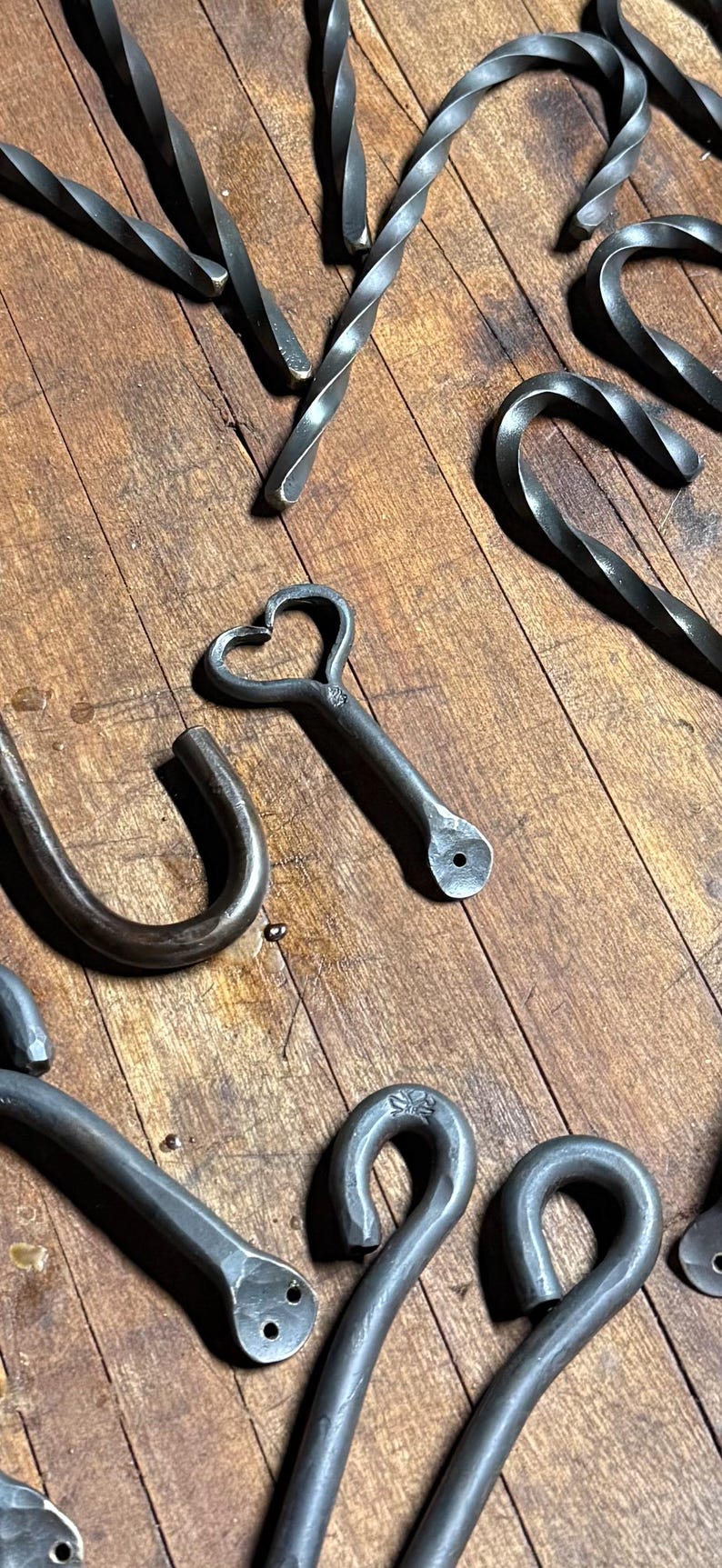 Op de afbeelding: Een verzameling donkergrijze, handgesmede metalen onderdelen, waaronder gedraaide staven, J-vormige haken en een hartvormig element. Verschillende onderdelen hebben platte uiteinden met voorgeboorde gaten voor montage en een ruwe afwerking.