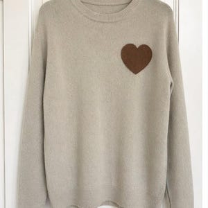 May include: A pale beige crew neck jumper featuring a brown heart motif on the left breast. The long-sleeved jumper is suspended from a black hanger against a white door.