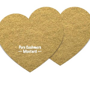 Può includere: Due toppe a forma di cuore di colore giallo senape, realizzate in morbido tessuto di cashmere. Il testo "Pure Cashmere - Mustard -" è stampato in bianco su uno dei cuori.