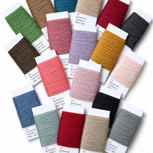 Könnte beinhalten: Eine Sammlung von einundzwanzig rechteckigen Karten, die mit verschiedenfarbigem 2-fädigem Garn umwickelt sind. Jede weiße Karte trägt den Aufdruck CC Reclaimed Cashmere und gibt Längen von 10m oder 20m an. Die Garnfarben umfassen Rot, Blau, Grün, Rosa, Gelb, Grau, Braun und Schwarz.