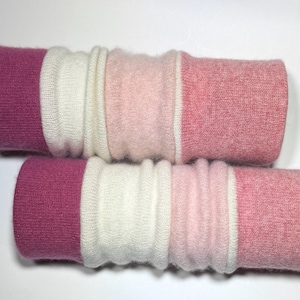 May include: A pair of knit leg warmers featuring a color-blocked design in shades of magenta, cream, light pink, and dusty rose. The fabric has a soft, textured appearance with ribbed sections and smooth bands arranged in a horizontal pattern.