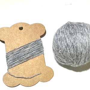 Puede incluir: Imagen artesanal con una bobina de hilo gris y una madeja de lana gris. El hilo está enrollado alrededor de una bobina de cartón marrón con borde festoneado. La madeja de lana es gris, esponjosa y texturizada.