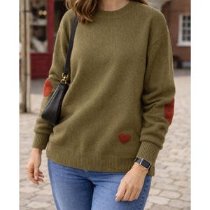 Op de afbeelding: Olijfgroene trui met rode hartvormige elleboogstukken en een klein hartdetail op de voorkant. De trui heeft een ronde hals en geribde manchetten. De persoon draagt een blauwe spijkerbroek en een zwarte handtas.