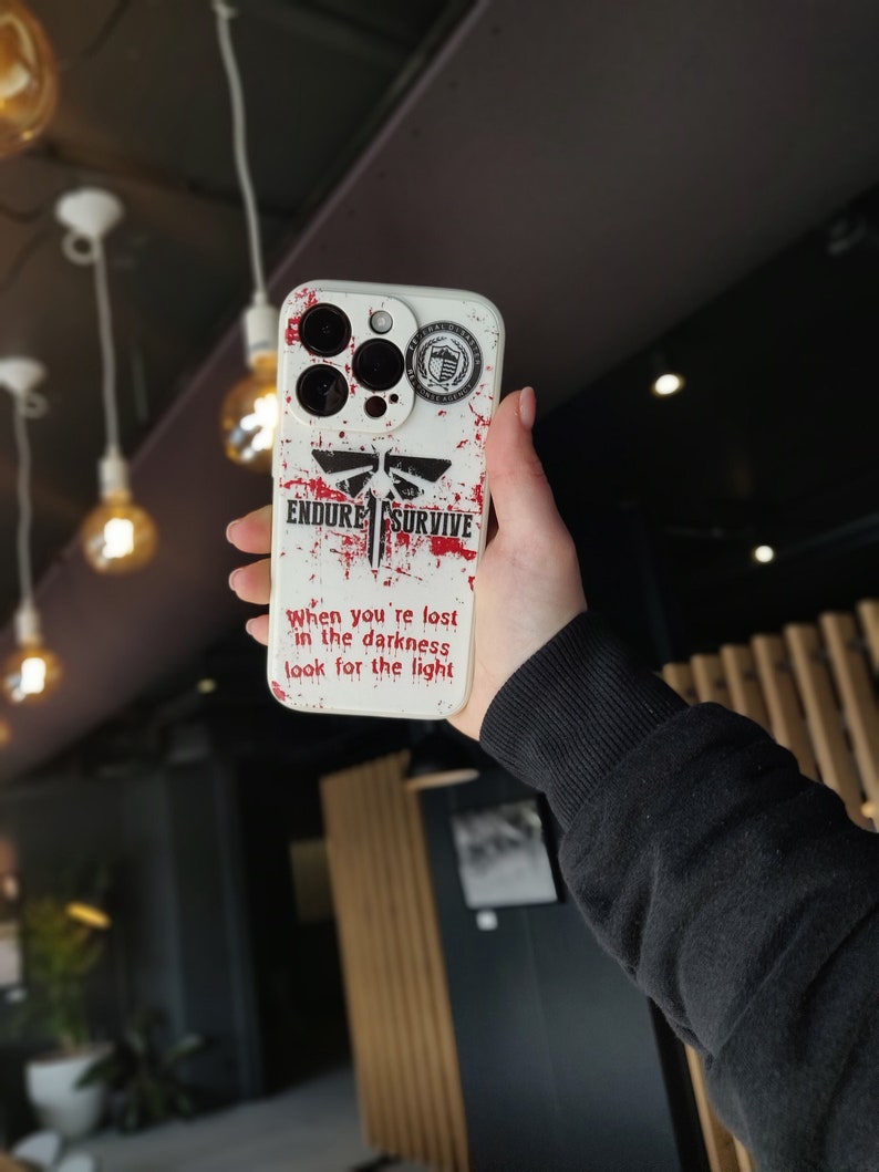 May include: White iPhone case featuring a red and black graphic of a dragonfly with the words "Endure" and "Survive". The case also has a small black and white sticker with a shield and the words "Federal Reserve" on it. The text "When you're lost in the darkness look for the light" is printed on the case.