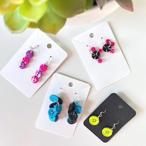 May include: Four pairs of earrings made from buttons. The earrings are displayed on white and black card stock. The earrings are made from pink, black, blue, and yellow buttons.