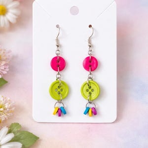 May include: A pair of handmade button earrings. Each earring features a hot pink button, a lime green button, and a cluster of colorful, small, rectangular charms. The earrings hang from silver-colored fish hook ear wires.