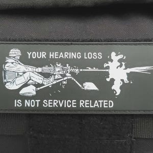 Puede incluir: Un parche rectangular negro con texto blanco y un gráfico de un soldado operando una ametralladora. El texto dice "YOUR HEARING LOSS IS NOT SERVICE RELATED". El parche está adherido a una mochila táctica negra.