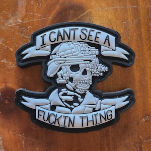 May include: A black and white patch featuring a skull wearing a helmet and night vision goggles. The text "I CAN'T SEE A" is above the skull, and "FUCKIN THING" is below.