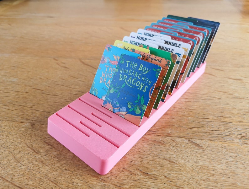 May include: A pink plastic card holder with slots for holding cards. The cards are all different colours and have titles like "The Boy Who Sang With Dragons" and "Horribile".