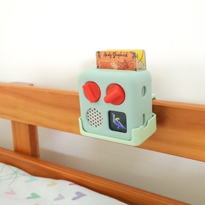 May include: A light green toy with red buttons and a small screen displaying a pixelated image of a dinosaur. The toy is attached to a wooden bed rail with a clip.