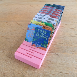 May include: A pink card holder with slots for holding cards. The holder is filled with cards featuring book covers with titles like "The Boy Who Sang With Dragons" and "The Human" and "Horrible Histories".