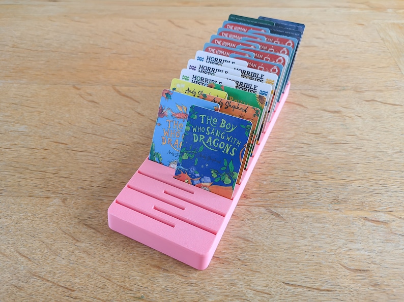 May include: A pink card holder with slots for holding cards. The holder is filled with cards featuring book covers with titles like "The Boy Who Sang With Dragons" and "The Human" and "Horrible Histories".