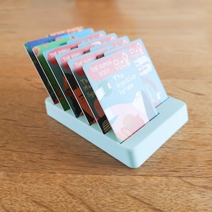 May include: A light blue card holder with a stack of colourful cards featuring illustrations and text about the human body. The cards include titles like "The Human Body", "The Digestive System", and "The Muscular System".