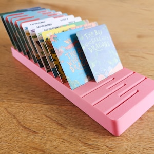 May include: Pink plastic card holder with slots for holding cards. The holder is angled and has a textured surface. The cards are visible and include book covers with titles like "The Boy Who Sang With Dragons".