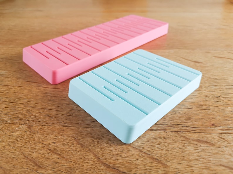 May include: Two rectangular soap dishes, one pink and one light blue, with raised lines on the surface.