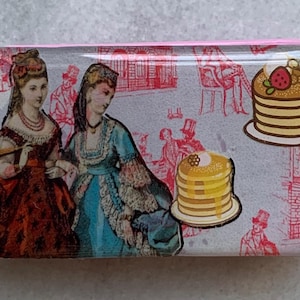 May include: A rectangular glass paperweight with a pink border. The paperweight features a vintage illustration of two women in dresses, a stack of pancakes with a strawberry on top, and a stack of pancakes with honey on top. The background is a light gray with a pink and red pattern.