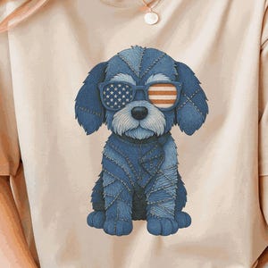 Peut inclure: T-shirt beige avec une illustration de chien bleu portant des lunettes de soleil avec un motif de drapeau américain. Le chien est fait d'un matériau semblable au denim. Le t-shirt est de coupe décontractée.