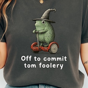 Peut inclure: T-shirt gris foncé avec une grenouille verte portant un chapeau de sorcière et conduisant un hoverboard rouge. Le texte "Off to commit tom foolery" est imprimé en dessous. Le mot "pepper" est imprimé en bas.