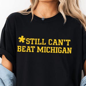 Peut inclure: T-shirt noir avec l'inscription "STILL CAN'T BEAT MICHIGAN" en jaune. Le t-shirt a un col rond et des manches courtes. La personne porte une veste en jean bleu clair par-dessus le t-shirt et un short en jean déchiré.