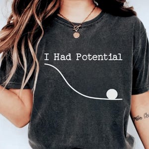Könnte beinhalten: Dunkelgraues T-Shirt mit dem Text "I Had Potential" über einer weißen Linienzeichnung, die einen Ball zeigt, der eine schräge Ebene hinunterrollt. Das T-Shirt hat einen lässigen Stil.