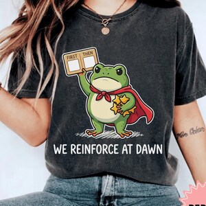 Könnte beinhalten: Ein anthrazitfarbenes Kurzarm-T-Shirt mit einer Grafik eines grünen Frosches, der einen roten Umhang trägt und ein Schild mit der Aufschrift FIRST THEN sowie gelbe Sterne hält. Der Text WE REINFORCE AT DAWN steht unter der Froschgrafik.