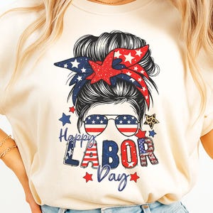 Könnte beinhalten: Elfenbeinfarbenes T-Shirt mit einem Grafikdesign zum Tag der Arbeit. Das Design zeigt ein Frauengesicht mit Sonnenbrille, einem rot-blauen Stirnband und dem Text "Happy Labor Day" in patriotischen Farben. Sterne und Streifen vervollständigen das Design.