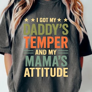Peut inclure: Un t-shirt gris foncé avec le texte "I GOT MY DADDY'S TEMPER AND MY MAMA'S ATTITUDE" en vert, orange et jaune. Le mot "pepper" est dans le coin inférieur gauche. Le t-shirt a une coupe décontractée.