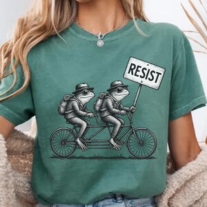 May include: Teal green t-shirt featuring a tandem bicycle with two frog illustrations wearing hats and backpacks. One frog holds a sign that reads "RESIST" in bold, white letters. The shirt is a casual style, suitable for everyday wear.
