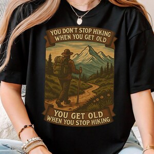 以下が含まれることがあります： ハイキングをテーマにしたグラフィックの黒いTシャツ。デザインは、バックパックとウォーキングスティックを持ったハイカーがトレイルを歩いている様子で、背景には山があります。テキストは「年をとってもハイキングをやめない、ハイキングをやめると年をとる」と書かれています。