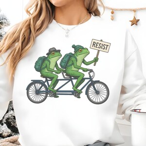 Peut inclure: Sweat-shirt blanc avec une illustration fantaisiste de deux grenouilles vertes sur un vélo tandem. L'une des grenouilles tient une pancarte avec le mot "RESIST". Le design comprend un vélo noir et des sacs à dos verts. Une tasse du Père Noël est tenue dans la main.
