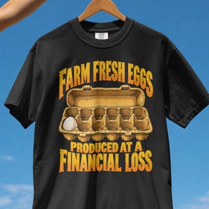 Könnte beinhalten: Ein schwarzes Kurzarm-T-Shirt mit einer Grafik eines offenen Eierkartons, der ein einzelnes weißes Ei enthält. Der Text darüber lautet FARM FRESH EGGS und der Text darunter lautet PRODUCED AT A FINANCIAL LOSS in fetter, gelber Schrift.
