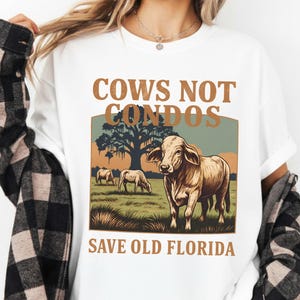 Peut inclure: T-shirt blanc avec un motif représentant une vache dans un champ, avec le texte "Cows Not Condos" et "Save Old Florida". Le design utilise des tons marron, vert et beige.