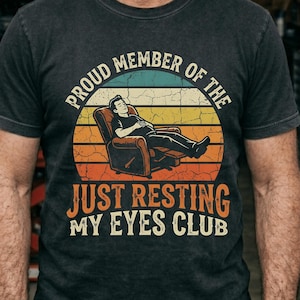 Peut inclure: T-shirt gris foncé avec un motif rétro d'une personne se relaxant dans un fauteuil, sur fond de coucher de soleil. Le texte sur le t-shirt se lit "PROUD MEMBER OF THE JUST RESTING MY EYES CLUB."