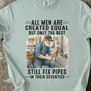 Könnte beinhalten: Hellblaues T-Shirt mit dem Text "ALL MEN ARE CREATED EQUAL BUT ONLY THE BEST STILL FIX PIPES - IN THEIR SEVENTIES". Die Grafik zeigt einen Klempner bei der Arbeit. Das Shirt ist aus weichem Material.