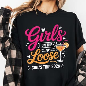 May include: Black t-shirt with the text "Girls on the Loose" in pink and orange, with a cocktail graphic. The shirt also includes "Girl's Trip 2026" in white. The person is wearing a black and brown plaid shirt.