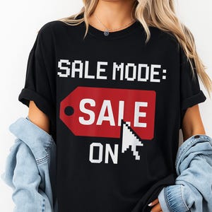 Peut inclure: T-shirt noir avec l'inscription "SALE MODE: SALE ON" en police pixelisée blanche et un graphique d'étiquette de prix rouge. Le t-shirt est de style décontracté, adapté au quotidien.