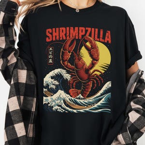 May include: Black t-shirt featuring a red lobster graphic emerging from ocean waves, with the text "SHRIMZILLA" in red. Japanese text is also included. The design is inspired by Japanese art.
