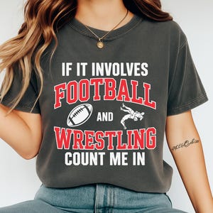Peut inclure: T-shirt gris foncé avec le texte "IF IT INVOLVES FOOTBALL AND WRESTLING COUNT ME IN". Le texte est en blanc et rouge. Un ballon de football blanc et une figurine de lutte sont également sur le t-shirt.