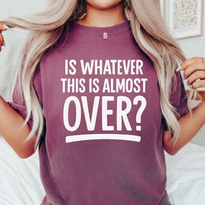 Pode incluir: T-shirt cor de baga com o texto branco "IS WHATEVER THIS IS ALMOST OVER?". A t-shirt tem mangas curtas e gola redonda. A palavra "BERRY" está impressa no canto inferior esquerdo.