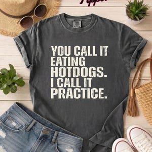 Peut inclure: T-shirt gris foncé avec le texte "YOU CALL IT EATING HOTDOGS. I CALL IT PRACTICE." L'image comprend un short en jean, des baskets blanches, un chapeau de paille et un sac fourre-tout tissé. Une petite plante en pot est également visible.