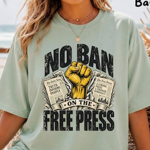 May include: A light green t-shirt featuring a graphic print with the text NO BAN ON THE FREE PRESS. The design includes a yellow raised fist centred between two newspapers, one labelled The Daily News and the other labelled The Free Press.